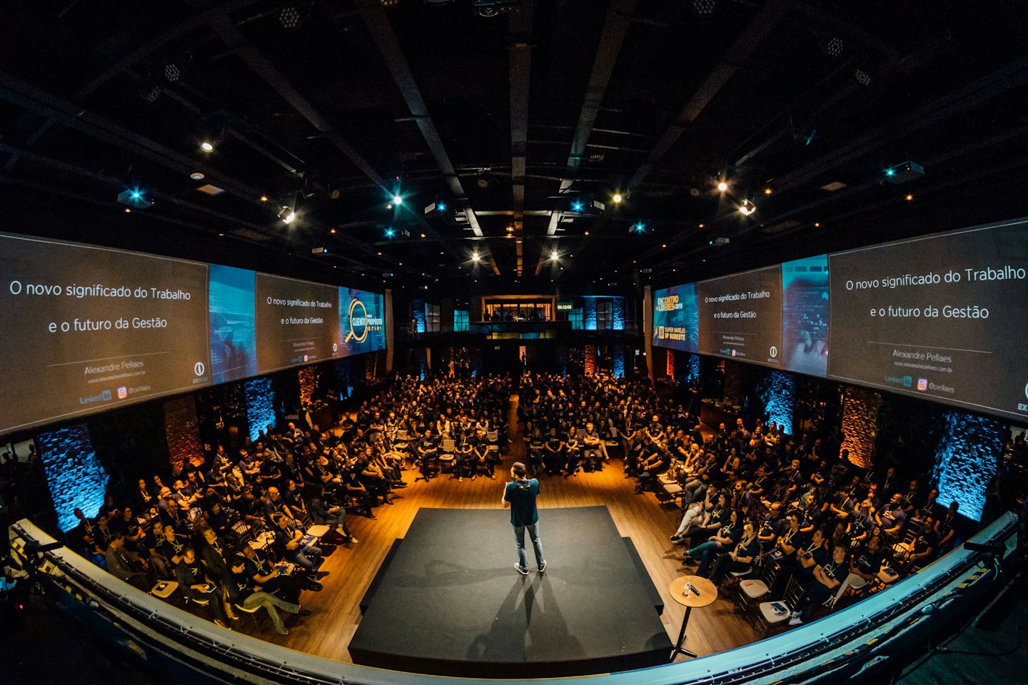 People attend a conference in a large, dark hall with a speaker on stage.