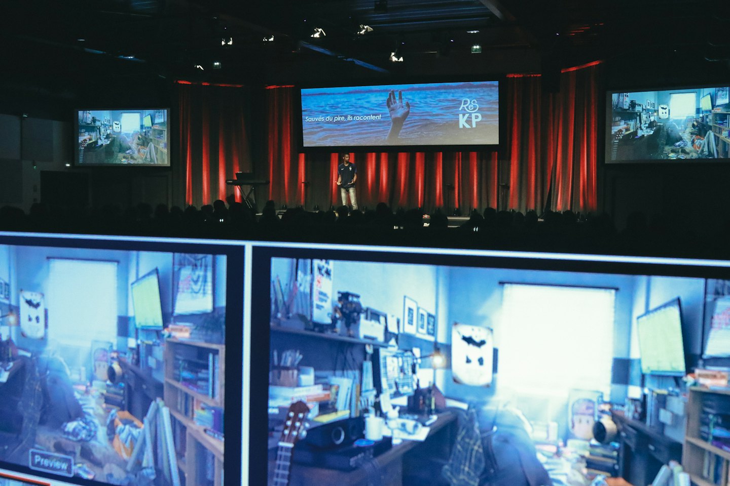 Speaker on a stage presenting to a large audience with screens.